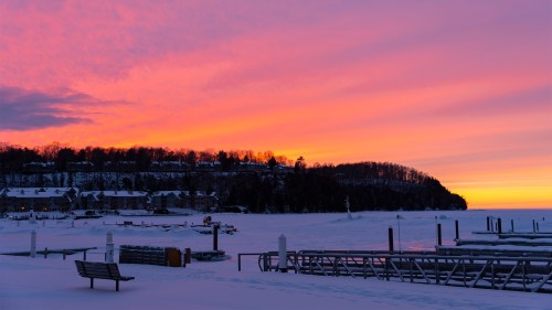 A serene winter sunset over a snowy lake, with colorful skies and silhouettes of trees and docks in the foreground.