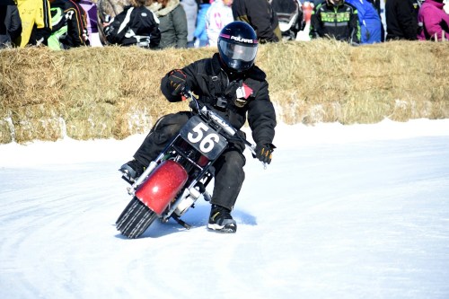 A rider in black gear races a small motorcycle on a snowy track, with spectators in the background.