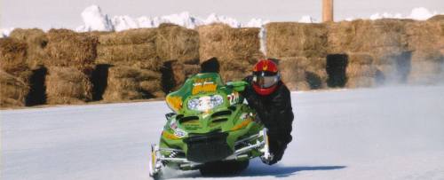 A snowmobiler leans into a turn on an icy track, with hay bales in the background and snow-covered ground.