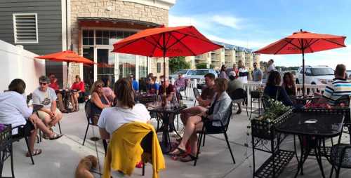 A lively outdoor café scene with people sitting at tables under orange umbrellas, enjoying food and drinks.