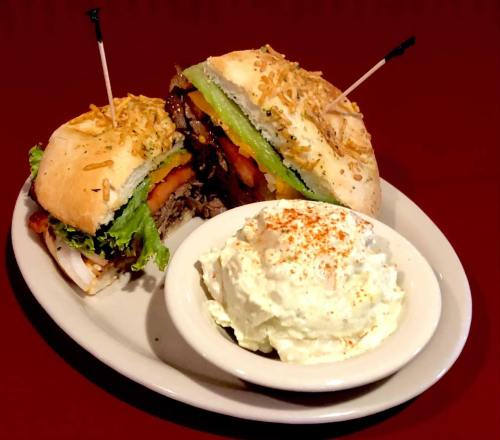 A plate featuring a large sandwich with layers of meat, vegetables, and cheese, alongside a bowl of creamy potato salad.