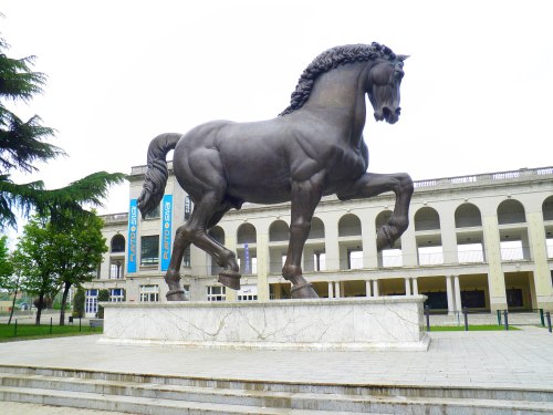 A large bronze horse statue stands on a marble pedestal in front of a building with arched windows.