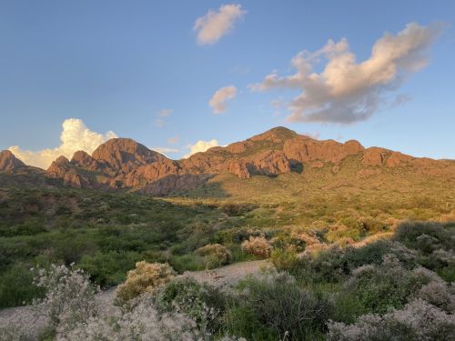 A scenic view of rugged mountains bathed in warm sunlight, surrounded by lush greenery and a blue sky with clouds.