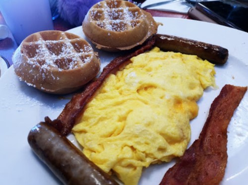 A plate with two waffles, scrambled eggs, sausage links, and crispy bacon, dusted with powdered sugar.