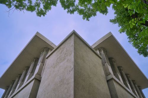 A low-angle view of a modern building with angular architecture, framed by green tree leaves against a cloudy sky.