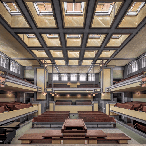 Interior of a spacious, modernist building with wooden benches, large windows, and a geometric ceiling design.