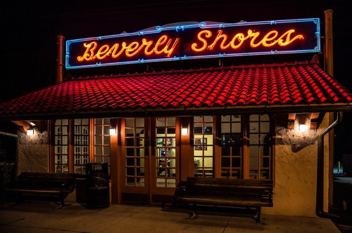Neon sign reading "Beverly Shores" above a warmly lit building with a tiled roof and benches outside at night.