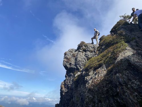 A person stands triumphantly on a rocky mountain peak, surrounded by clouds and blue sky.