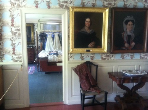 A historic room with patterned wallpaper, portraits on the walls, and a view into a bedroom with vintage furnishings.