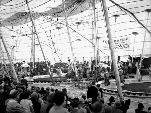 A vintage circus scene inside a large tent, with performers and a captivated audience watching the show.