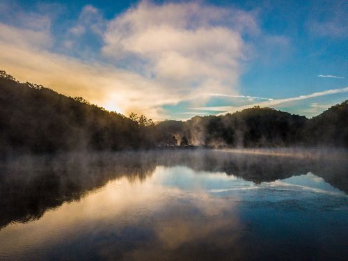 A serene lake at sunrise, surrounded by misty hills and a colorful sky reflecting on the water's surface.