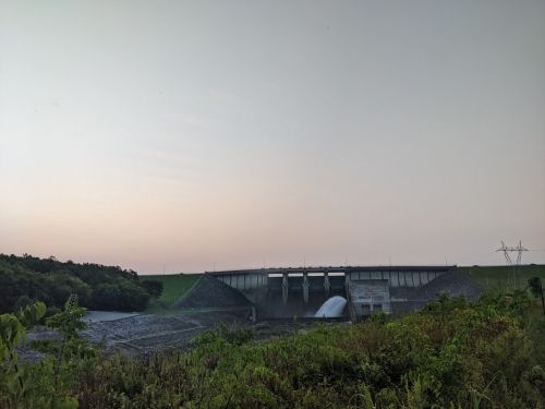 A dam with water flowing from its gates, surrounded by greenery and a clear sky at dusk.