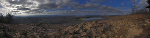 A panoramic view of a rocky landscape overlooking a valley with a river and fields under a cloudy sky.