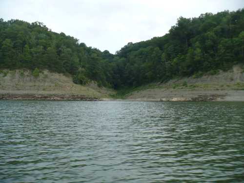 A calm lake surrounded by green hills and trees, with a narrow inlet visible in the distance.