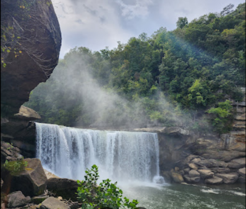 A serene waterfall cascades into a river, surrounded by lush greenery and mist rising from the water.