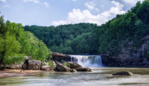 A serene waterfall cascades into a calm river, surrounded by lush green trees and a clear blue sky.