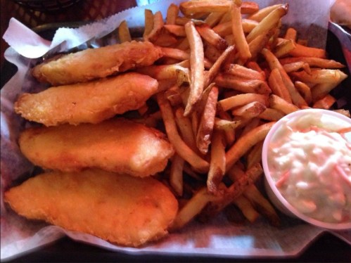 A plate of golden fried fish fillets served with crispy French fries and a side of coleslaw in a small cup.