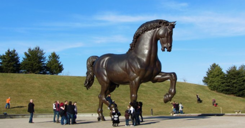 A large bronze horse statue stands tall, with people gathered around it in a park under a clear blue sky.