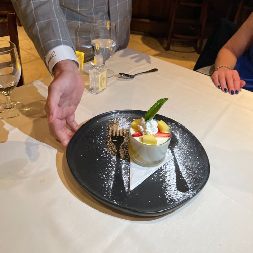 A waiter serves a dessert topped with fruit and whipped cream on a black plate, with a hand reaching out to present it.