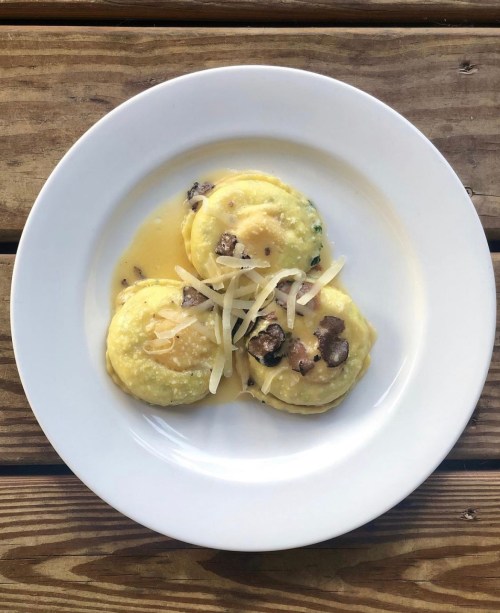 Three ravioli topped with cheese and mushrooms, served in a light sauce on a white plate, set on a wooden table.