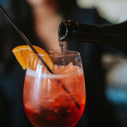 A close-up of a cocktail being poured into a glass with ice and an orange slice garnish.