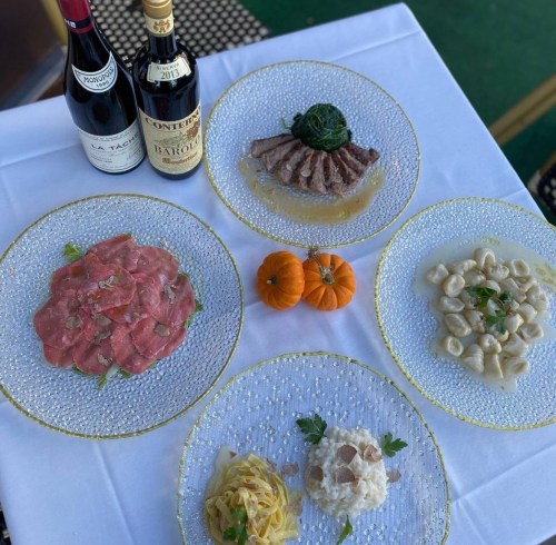 A table set with four elegant plates of Italian dishes, two bottles of wine, and small pumpkins for decoration.