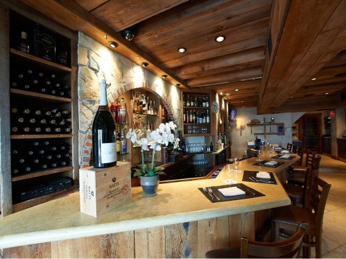 A cozy bar with wooden beams, stone accents, and shelves filled with wine bottles and spirits, featuring a floral centerpiece.