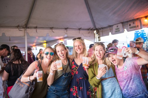 A group of five women smiling and holding drinks under a tent at a lively outdoor event.