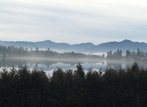 A serene lake surrounded by misty mountains and trees, reflecting a calm, overcast sky.