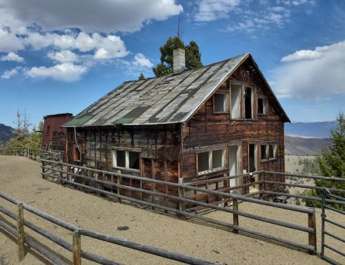 A weathered wooden cabin with a slanted roof, surrounded by a wooden fence and set against a blue sky with clouds.