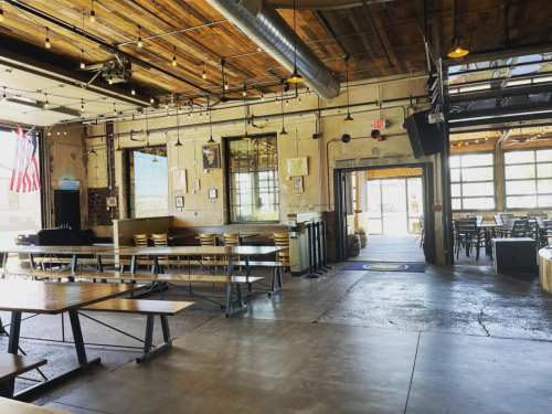 Interior of a spacious restaurant with wooden beams, long tables, and large windows, featuring a casual atmosphere.