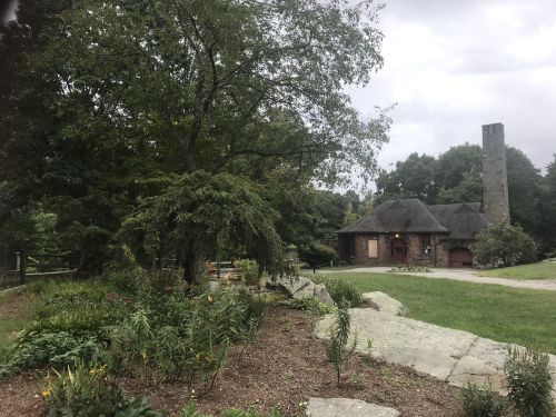 A stone building with a thatched roof surrounded by greenery and a garden, under a cloudy sky.