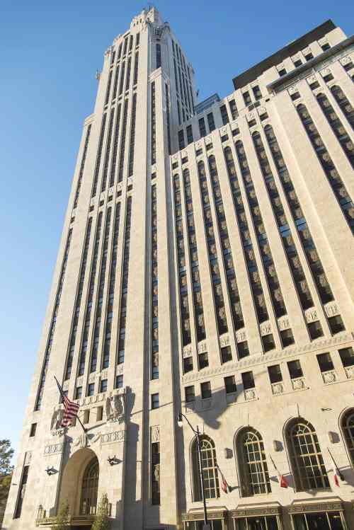 A tall, Art Deco-style skyscraper with a blue sky backdrop, featuring flags and decorative stonework at the entrance.