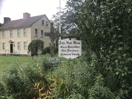 Sign for John Hunt House, home of the East Providence Historical Society, surrounded by greenery. Historic building in background.