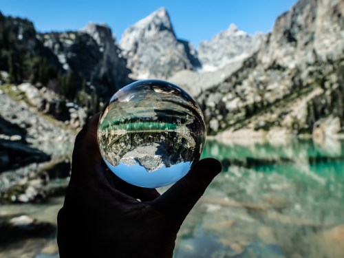 A hand holds a crystal ball reflecting a mountain landscape and turquoise lake under a clear blue sky.