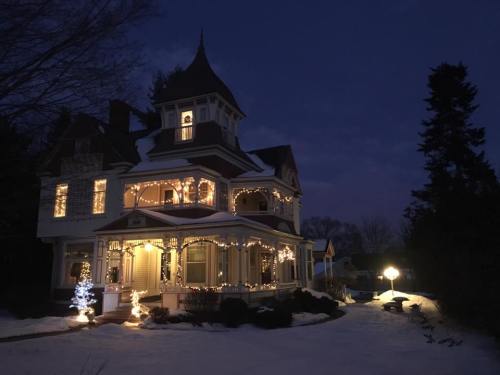 A beautifully lit Victorian house at dusk, adorned with holiday lights and surrounded by snow.