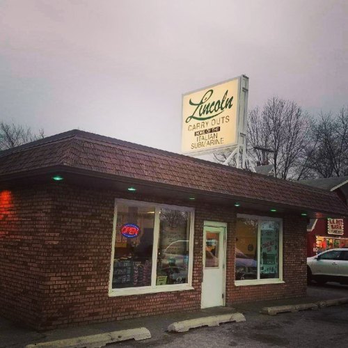 A brick building with a sign reading "Lincoln Carry Outs" and a window displaying an open sign, set against a cloudy sky.