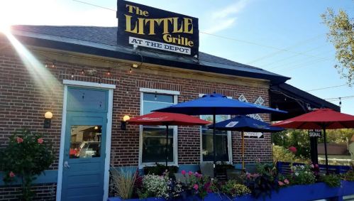 A cozy restaurant with a sign reading "The Little Grille at the Depot," featuring colorful umbrellas and flower planters.