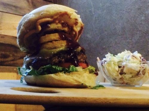 A towering burger with lettuce, pickles, and sauce, served with a side of coleslaw on a wooden board.