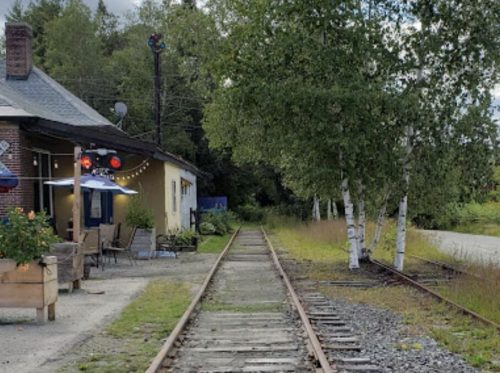 A view of a railway track leading towards a building, surrounded by trees and a cloudy sky.