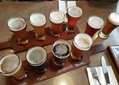A flight of ten different craft beers in small glasses on a wooden tray, with labels for each type.