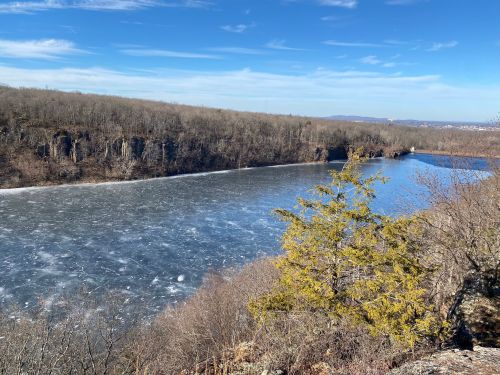 A scenic view of a frozen river surrounded by bare trees and rocky cliffs under a clear blue sky.