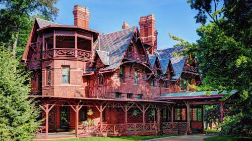 A large, ornate red brick mansion with multiple gables and chimneys, surrounded by lush greenery and blue skies.