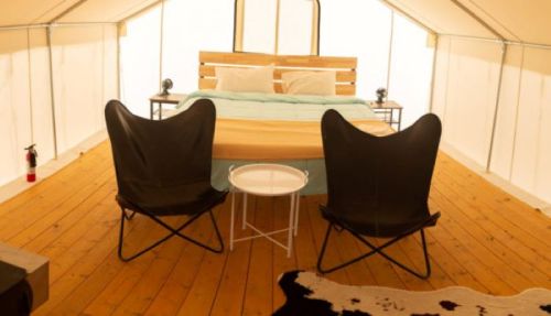 Cozy glamping tent interior with a bed, two chairs, and a small table on wooden flooring.