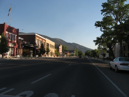 A quiet street scene with buildings, trees, and mountains in the background under a clear blue sky.