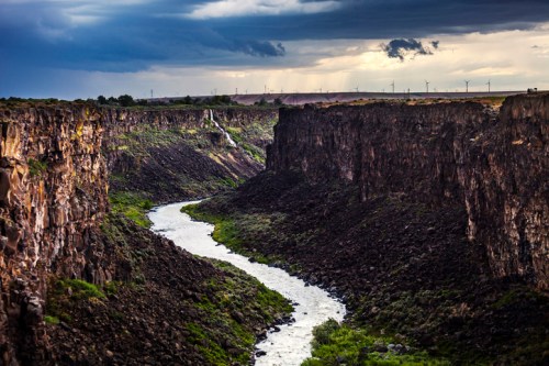 A winding river flows through a rocky canyon under a cloudy sky, with wind turbines visible in the distance.