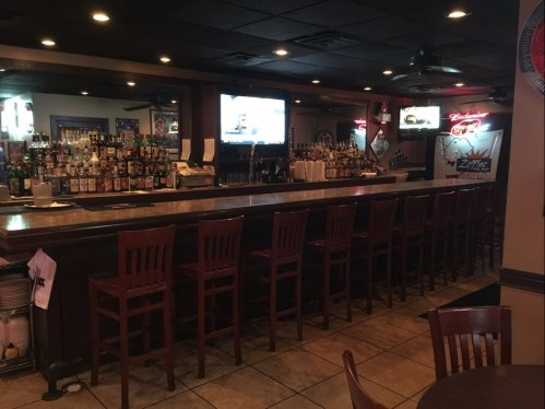 A dimly lit bar with a long counter, high stools, shelves of liquor, and a TV displaying a sports game.