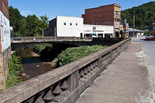 A stone bridge over a small creek, with buildings and trees in the background under a clear blue sky.