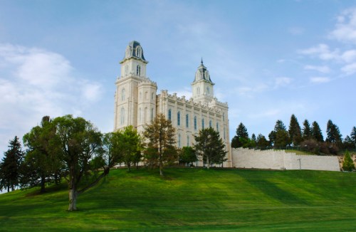 A grand white temple on a green hillside, surrounded by trees under a blue sky with scattered clouds.
