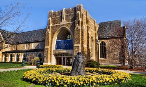 Historic stone building with a large arched entrance, surrounded by vibrant yellow flowers and clear blue sky.